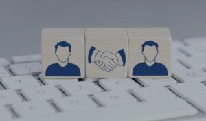Picture2 - RQTech - RQTech Two user icons on either side with a handshake symbol between them on wooden blocks, set on a keyboard background (teamwork/partnership theme).