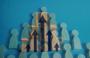 Group of wooden human shapes indicating growth surrounding a central stack of blocks with blue upward arrows, symbolizing teamwork and growth on a blue background.