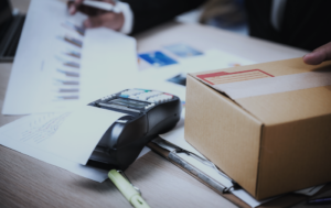 Cardboard shipping box on a desk beside a receipt printer and scattered papers on a busy workspace.